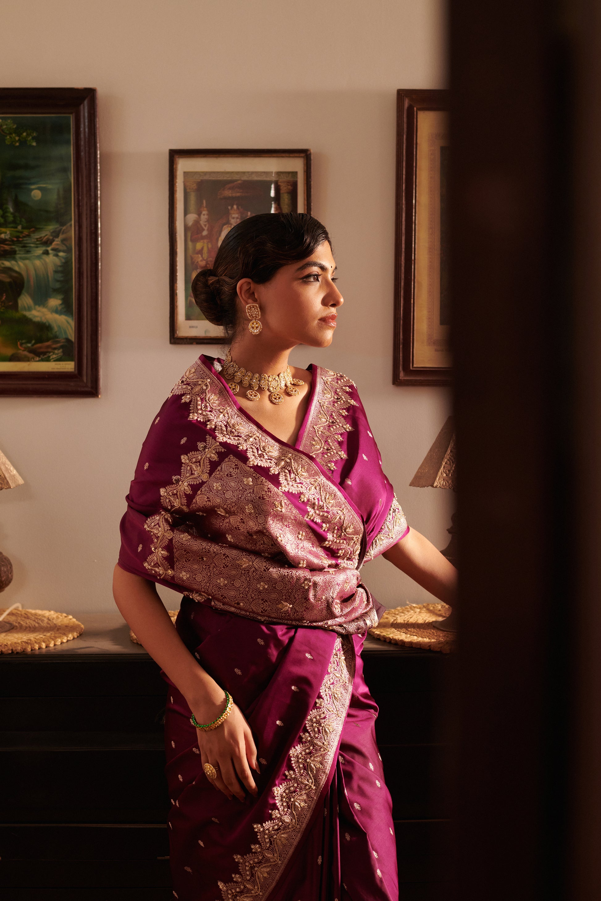Woman in a traditional purple and gold saree standing in a room with framed pictures on the wall.